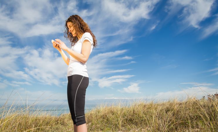 Woman in t-shirt and athletic gear checking phone with coast in background